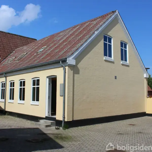 Et gult hus med rødt tegltag står stille i en gårdsplads omgivet af en mur og et metalgitterhegn. Himlen er blå med få hvide skyer.