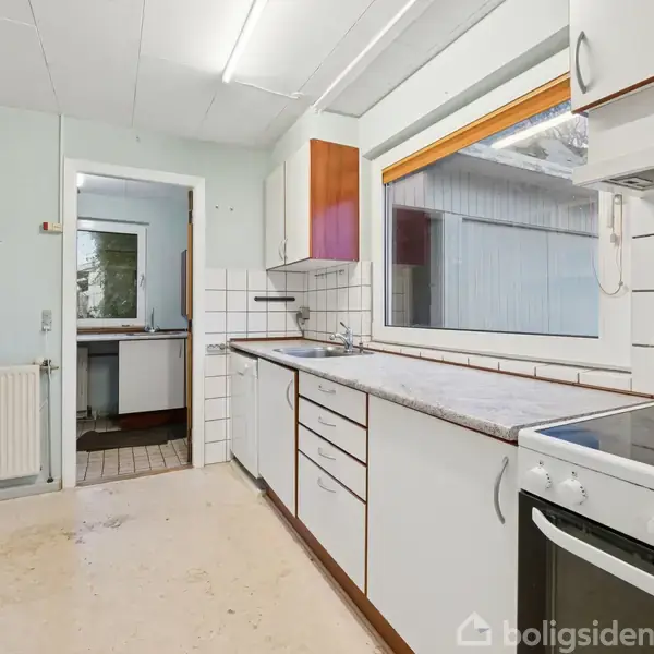 Et køkken med hvide skabe og skuffer langs væggen. Et komfur står til højre, mens et stort vindue giver udsigt til en overdækket terrasse. Væggene er lyseblå.