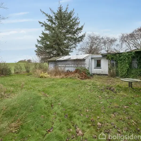 Græsplæne omgivet af et hus med hvid facade dækket af klatreplanter. Til højre står et rundt bord. Baggrunden er præget af store træer og en åben mark.