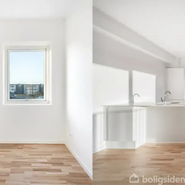 En lys stue med trægulv og et vindue, der viser udsigt over bygningskomplekser. Til højre en moderne, hvidt køkken med indbyggede skabe og vask på køkkenbordet.