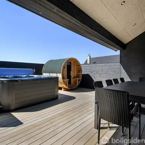 Udendørs terrasse med træbord og sorte stole. Et boblebad står til venstre, og en rund sauna er bagved. Omgivelserne er moderne og solrige.