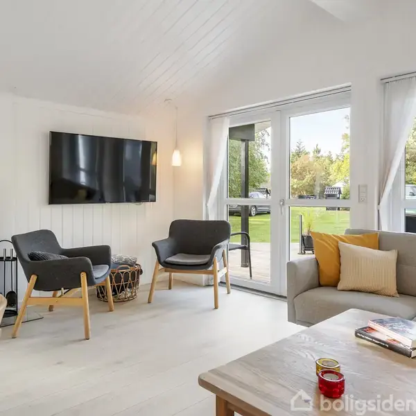 Stue med sofa, lænestole og et sofabord med bøger og kaffekrus. En fladskærm hænger på væggen ved siden af en brændeovn. Store vinduer viser en grøn have udenfor.