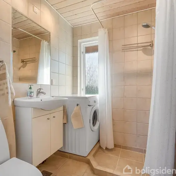 Et badeværelse med toilet og håndvask. En vaskemaskine står ved siden af vasken. Bruseområdet har et hvidt forhæng. Vægge og gulv er flisebelagt, loftet i lyst træ.
