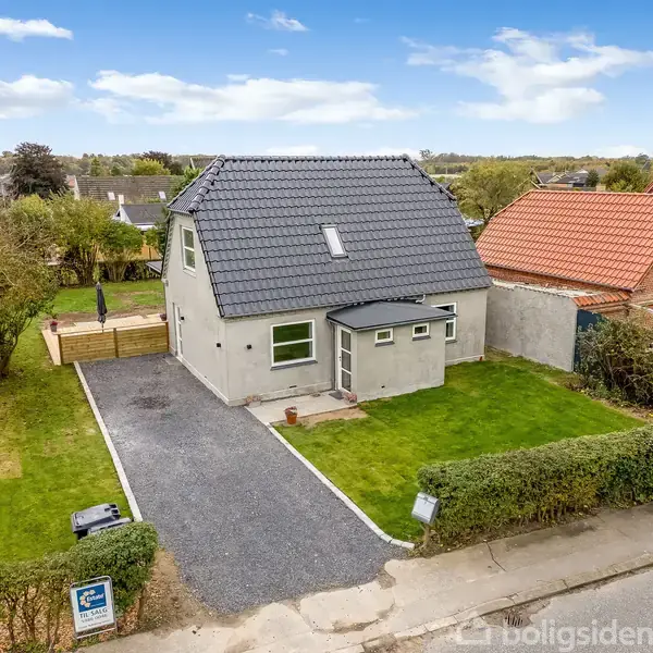 En to-etagers gråmalet hus med sort tag står i en grøn have. En grusbelagt indkørsel fører til hovedindgangen. En anden bygning ses ved siden af, omgivet af hække og træer.