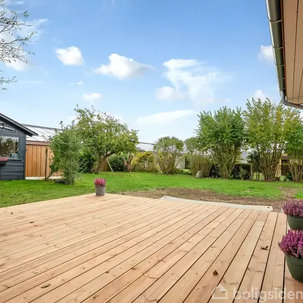 Terrasse lavet af lyse brædder med blomsterpotter, beliggende ved et sortmalet hus. I baggrunden ses en have med grønne buske og et skur, under en blå himmel.