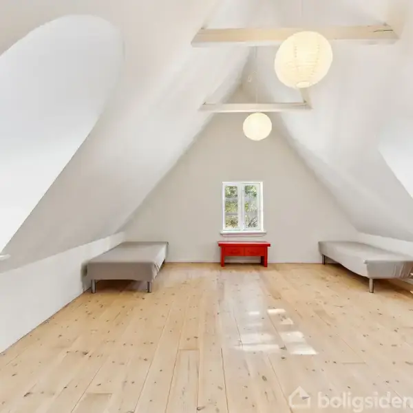 Loftværelse med to senge placeret langs hver side. Et rødt bord står ved gavlvæggen under et lille vindue. Rummet er lyst med trægulv og hvide vægge.