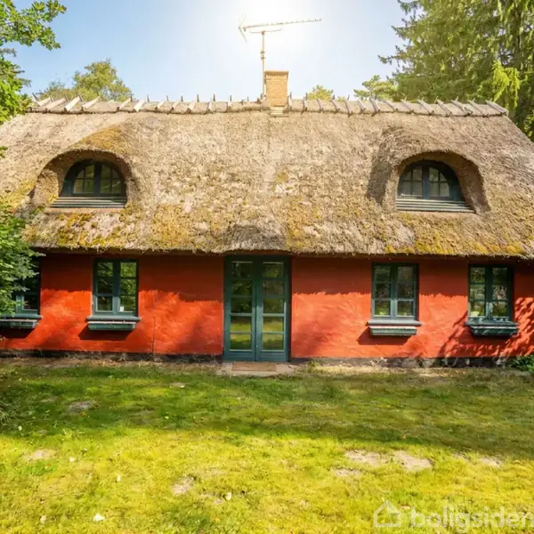 Rødt hus med stråtag og sprossede vinduer står i en grøn have omgivet af træer. Sollys skinner på taget, og der er en græsplæne foran huset.