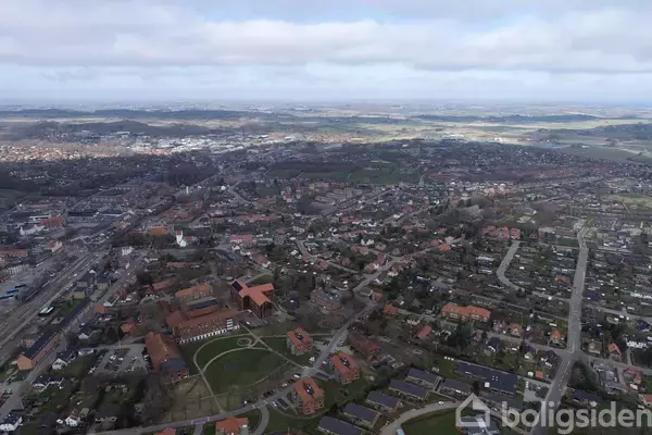 Der er et luftfoto af en by med mange huse og veje, omgivet af åbne grønne områder og marker i horisonten under en overskyet himmel.