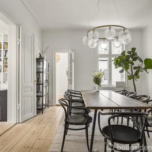 Langt spisebord med sorte stole omkring, placeret i et lyst rum med trægulv. En moderne lysekrone hænger over bordet. Store vinduer og grønne planter skaber en indbydende atmosfære.