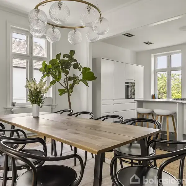Et spisebord med sorte stole står i et lyst køkken-alrum. Der er en stor plante og en vase med blomster på bordet. Køkkenet har hvide skabe og en kogeø.