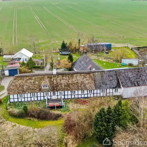 Bindingsværkshus med stråtag omgivet af have. Huset ligger på en gård med flere mindre bygninger. I baggrunden ses store, åbne marker.