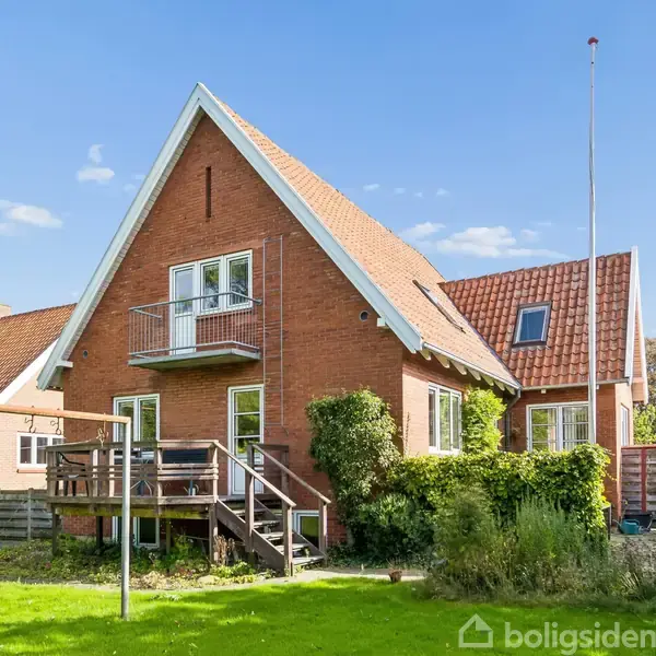 Rødt murstenshus med balkon og trappe forbundet til en hævet terrasse. I haven ses græsplæne, en flagstang og hvid plasticstol. Skyfri himmel og nabohus i baggrunden.