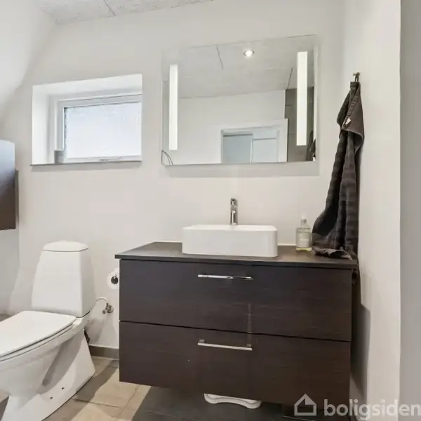 Toilet og vask er placeret mod en hvid væg i et badeværelse med moderne indretning. Over vasken hænger et spejl og et håndklæde, mens brusebadet er til højre.
