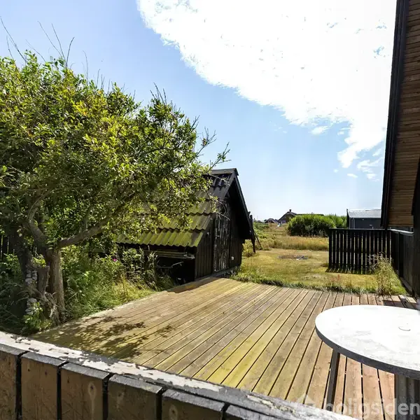 Et rundt bord står stille på en træterrasse ved siden af et sortmalet hus. En frodig have med et træ omkranser området under en klar blå himmel.