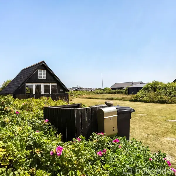 En sort træbygning står stille ved siden af en græsplæne omgivet af buske med lyserøde blomster. En postkasse med nummeret "21" ses i forgrunden.