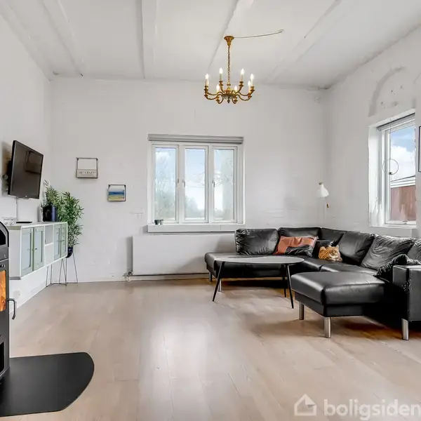 En sort brændeovn opvarmer stuen, hvor en sort lædersofa står foran store vinduer. Væggene er hvide, og en lysekrone hænger fra loftet. En vægmonteret TV ses til venstre.