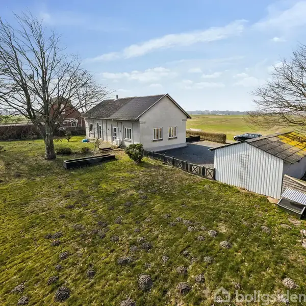 Et hvidt hus står stille omgivet af en rummelig have med græs og træer. Ved siden af huset er en hvid garage i et åbent, landligt landskab.