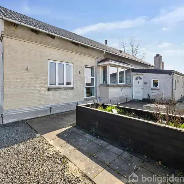Et hvidt murstenshus med sort tag står i en solrig have med flisebelagt gangsti og små bede. Huset har flere vinduer og en veranda mod venstre.