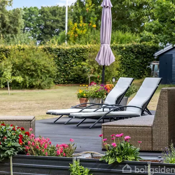 To liggestole står på en terrasse omgivet af blomster i farverige krukker. En parasol er rejst mellem stolene. Græsplænen og buske ses i baggrunden.
