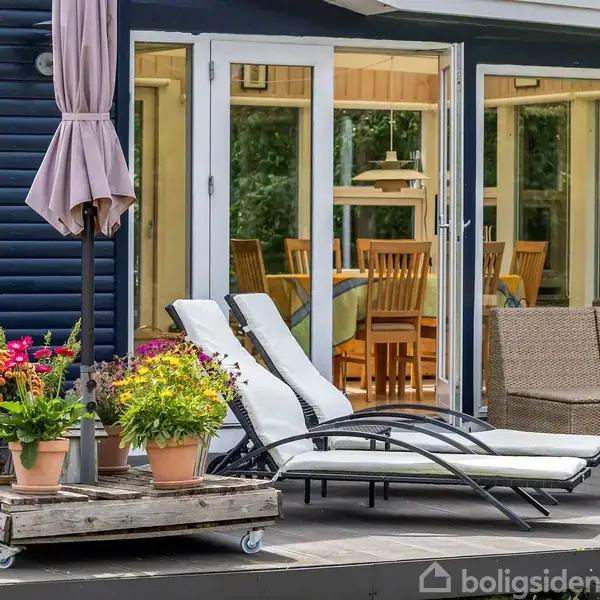 To liggestole står på en træterrasse foran et hus med store vinduer. En parasol er foldet sammen, og blomster i potter står ved siden af liggestolene.