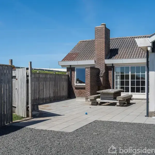 Hus med rød murstensskorsten står på en flisebelagt terrasse med gråt grus. Et træbord med bænke er placeret foran. En træhegn omgiver området under en blå himmel.
