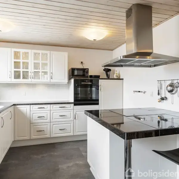 Køkken med hvide skabe og skuffer, sort kogeø med emhætte. Ovenover er der et trælignende loft og en rund loftslampe. Væggen til højre har hængende køkkenredskaber.