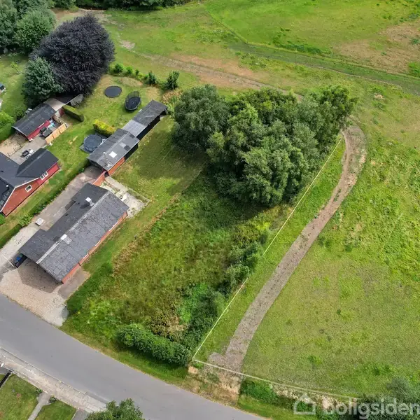 Parcelhusområde med flere røde teglhuse og haver, placeret langs en vej. Til højre en stor grøn mark med en buet grussti, omkranset af træer. Ingen tekst synlig.