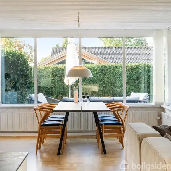 Et spisebord med stole står i en lys spisestue med store vinduer. Udenfor er der hække og en del af et hus. På bordet hænger en hvid lampe.