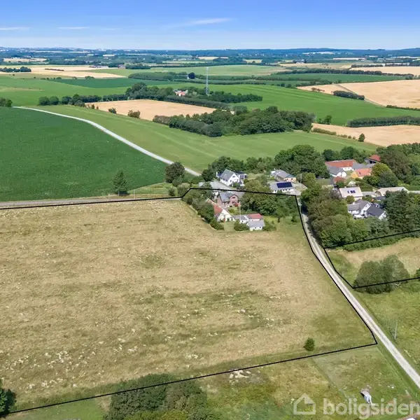 Landsby omgivet af marker; huse danner en lille klynge langs veje. Grønt og gyldent landskab strækker sig til horisonten under en klar blå himmel.