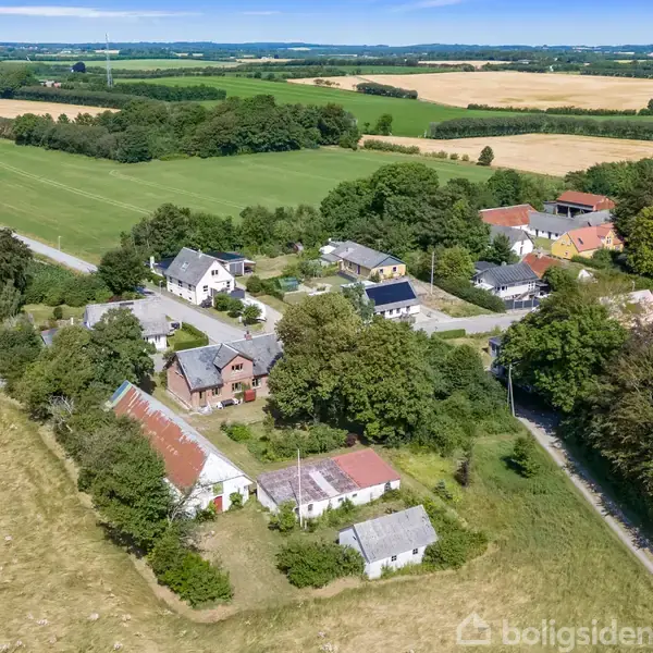 Landsby med spredte huse omgivet af marker og træer. Bygningerne er placeret langs snoede veje, mens det åbne landskab strækker sig mod horisonten under en klar himmel.