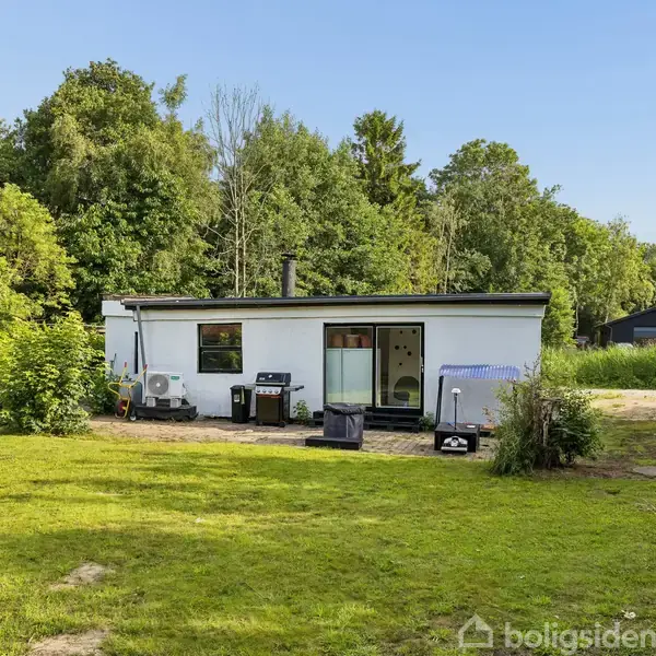 En lille, hvid bygning med fladt tag står omgivet af en græsplæne og tæt skov. En terrasse foran huset har en grill og nogle stole.