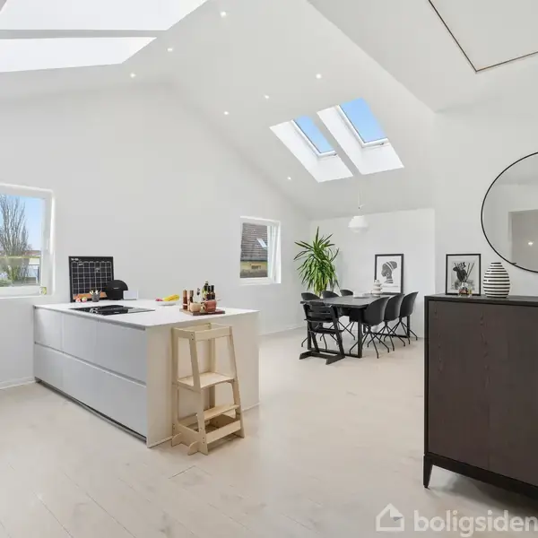 Et moderne køkken med en stor hvid ø, to barstole og et vindue. Ved siden af er et spisebord med stole. Loftet har ovenlys og rummet er lyst og rummeligt.