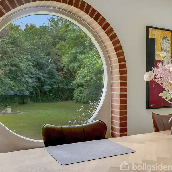 Et rundt vindue indrammet af mursten viser en grøn have med træer. Foran vinduet står et spisebord med blomster i en vase. En maleri hænger på væggen.