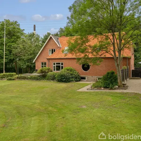 Et rødt murstenshus med rødt tegltag står i en frodig have fyldt med græs og træer. En flisebelagt sti fører forbi huset.