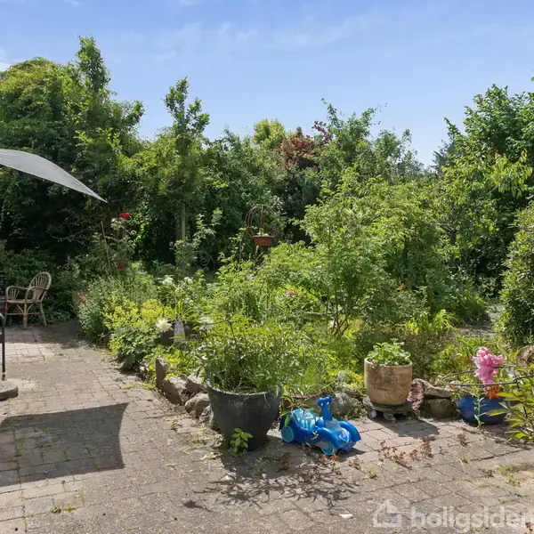 En brostensbelagt terrasse med havemøbler og en parasol står i forgrunden. Omgivet af frodige buske og træer i en solrig have, ses en blå plastiklegeting og flere potteplanter.