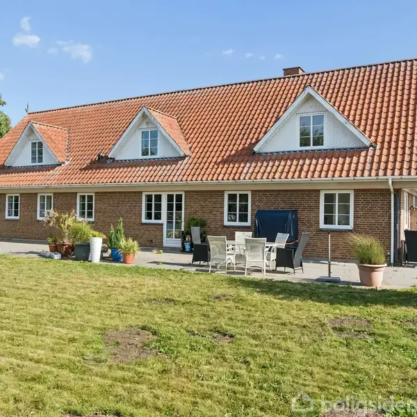 Et rødt murstenshus med fire gavlvinduer og teglstenstag står i en have. Foran huset er en terrasse med havemøbler og krukker med planter. I baggrunden ses grønne træer.