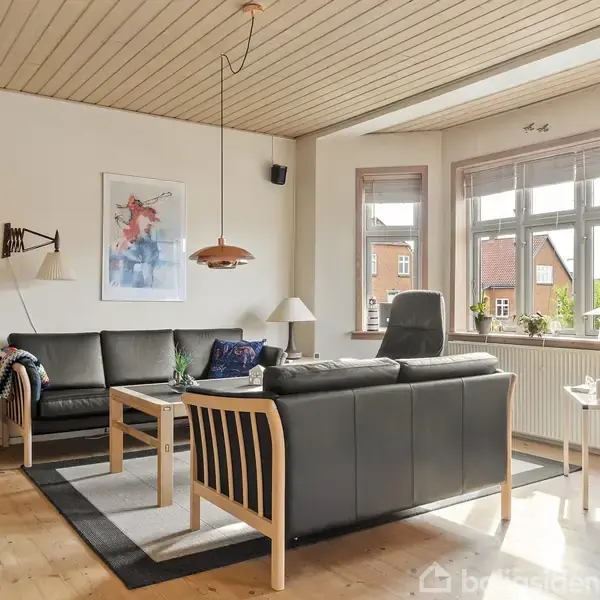 En lys stue med sorte lædersofaer omkring et træ-kaffebord, dekorativ plakat på væggen, store vinduer med udsigt til røde murstenshuse, lyse trægulve og loftslamper.