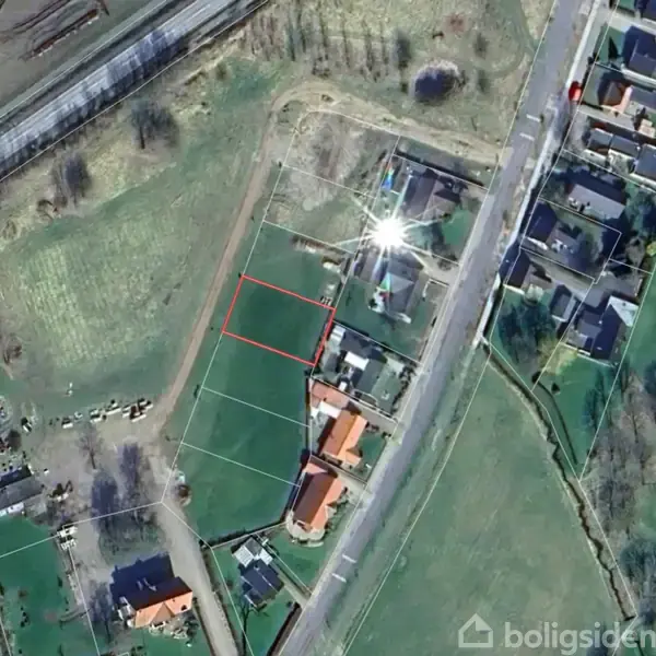 En by set fra luften med en sø i baggrunden. Huse og gårde spredt over grønne marker. En vej løber gennem landskabet. Skyer svæver på himlen.