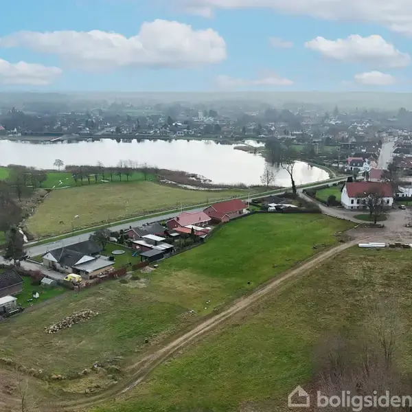 Landskab med grønne marker i forgrunden, små huse og bygninger langs en vej i midten, og en stor sø omgivet af bebyggelse i baggrunden under en blå skyet himmel.