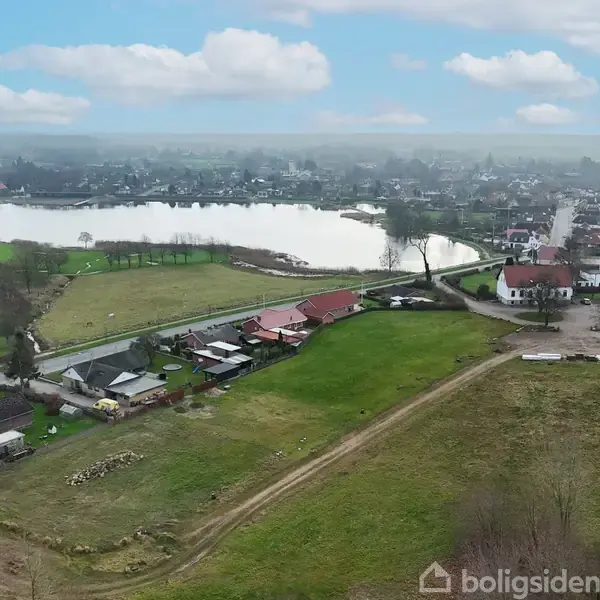 Landskab med grønne marker og spredte huse ved en sø. I baggrunden ses en by, og over himmelen er der spredte skyer.