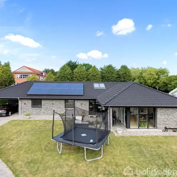 Et moderne enfamiliehus med solcellepaneler på taget. En trampolin står på græsplænen i haven foran huset. Til venstre holder en bil i indkørslen.