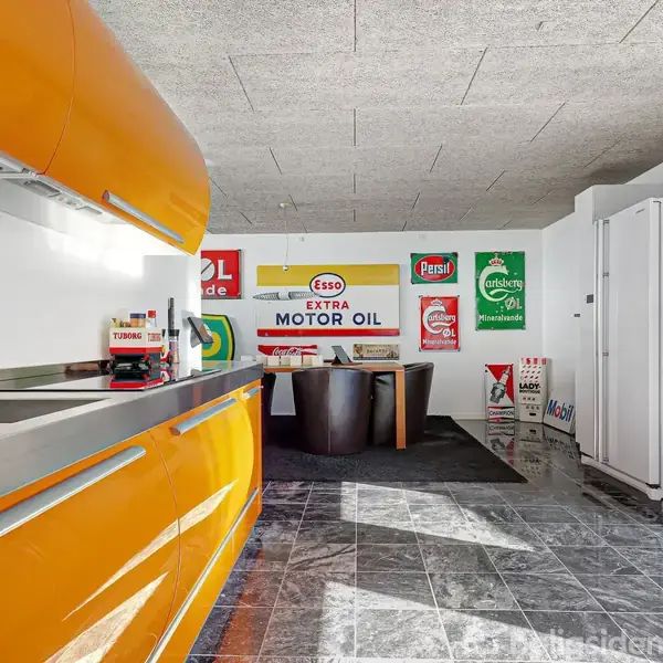 En orange, moderne køkkenø med vask i forgrunden står i et rum med vintage emaljeskilte på væggene. Gulvet er belagt med grå fliser, og der er hvide skabe.