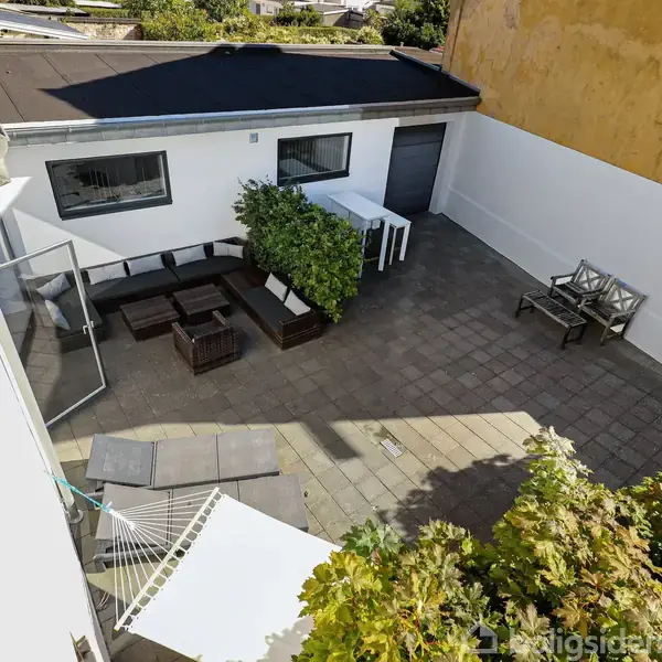 Terrasse med sofaarrangement af mørkt træ og puder i hjørnet. Hvide borde og grønne planter står tæt ved. Fliser dækker området, omgivet af hvide og gule vægge.