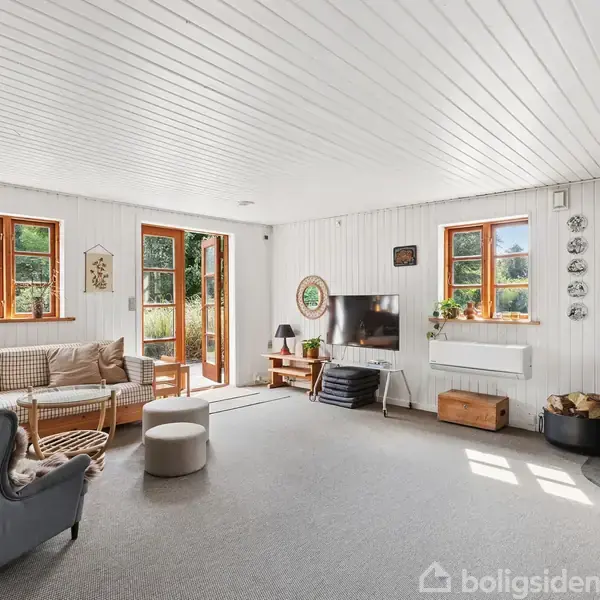 Stue med brændeovn, sofa og lænestol, der skaber en hyggelig atmosfære. Store vinduer og dør åbner ud mod en grøn have og slipper sollys ind. Fjernsyn og dekorationer ses.