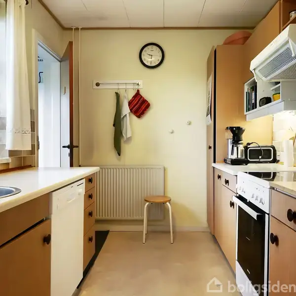 Køkken med træskabe og hvidevarer. En stol står ved radiatoren under et vægur. Køkkenredskaber hænger på væggen. Der er et vindue med blondegardin og planter.
