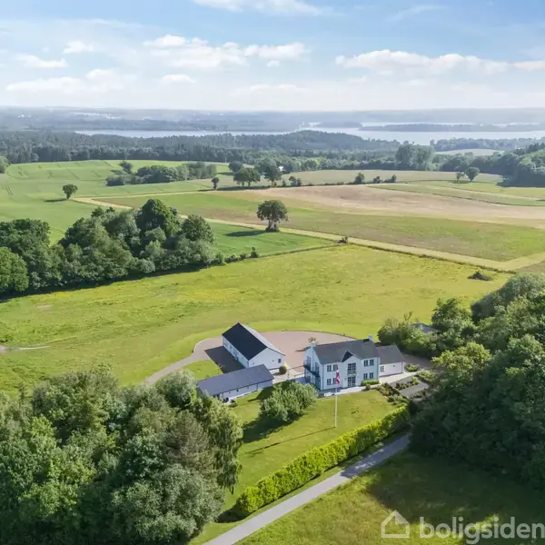Hus omgivet af marker ligger i naturskønt landskab med skov og søer i baggrunden. Blå himmel og sollys skaber en fredfyldt atmosfære.