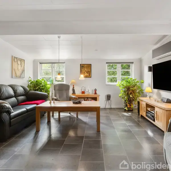Sofa og lænestol står rundt om et træstuebord i en lys stue. Vægge har kunstmalerier, mens et TV er monteret over en skænk. Grønne planter står ved vinduerne.