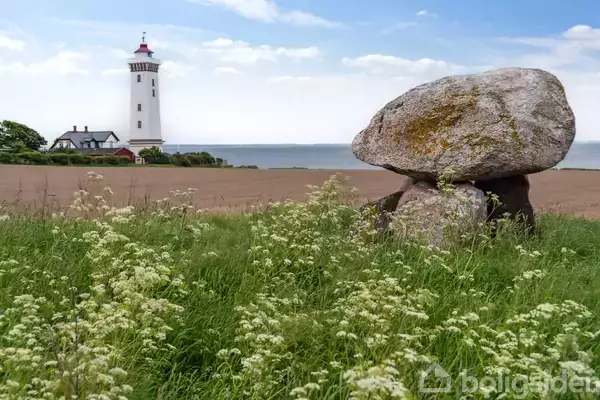 En stor sten står stille i en mark med græs og blomster; i baggrunden ses et hvidt fyrtårn nær kysten under en klar blå himmel.