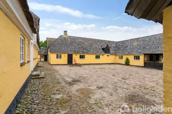 En gul bindingsværk bygning omkranser en brostensbelagt gårdsplads. Bygningen har et stråtækt tag, og man kan se grønne træer i baggrunden under en blå himmel.