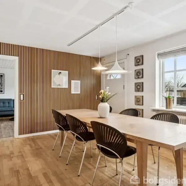 Spisebord med seks stole står i en lys spisestue. En vase med blomster står på bordet. Væggene har træpaneler og vinduerne slipper naturligt lys ind.